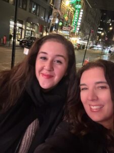 Two women smiling for a selfie on a city street at night, capturing the warmth of connectedness amid bright storefront lights, a neon pizza sign, and the lively backdrop of cars and city buildings.