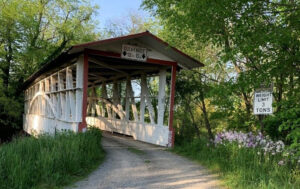 A rustic white covered bridge with red trim stands over a gravel road, surrounded by green trees and grass. Signs spark curiosity about its 12-foot clearance and 3-ton limit, while wildflowers bloom along the roadside.