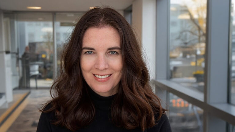 Lynn A. Haller, a woman with long brown hair and blue eyes, smiles at the camera. She is wearing a black turtleneck and is indoors with a softly blurred background.