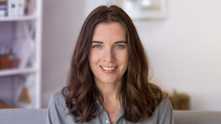 Lynn A. Haller, a woman with long brown hair and blue eyes, smiles at the camera. She is wearing a gray button-up shirt and is indoors with a softly blurred background.