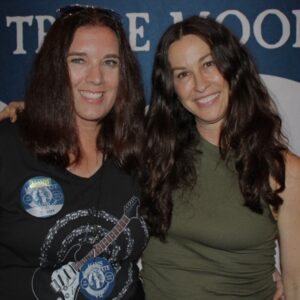 Two women with long brown hair stand side by side, smiling at the camera. One wears a black shirt with a guitar graphic and event badges, while the other wears a sleeveless olive-green top. A blue and white backdrop is behind them.