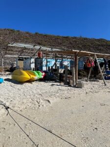 A small open-air kayak rental shelter on the sandy beach at Playa Pichilingue, with colorful yellow and green kayaks stacked in the foreground and a rustic wooden structure behind them.