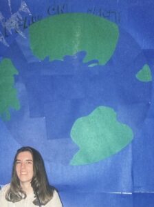 Lynn A. Haller sitting at a staff desk in front of a large handmade globe display made from blue and green tissue paper, created for a Christmas Around the World decorating contest at a residential youth program.